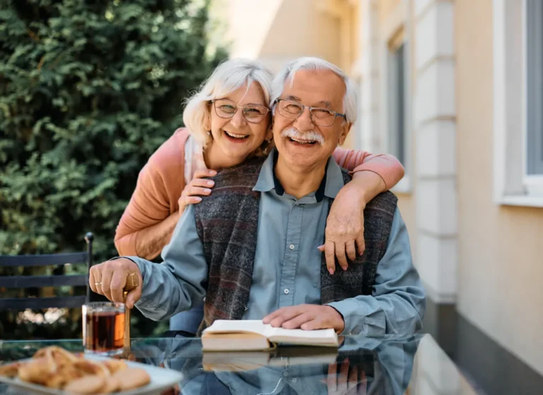 Smiling older couple sitting at an outdoor table with a book and drink, embracing and enjoying each other's company.