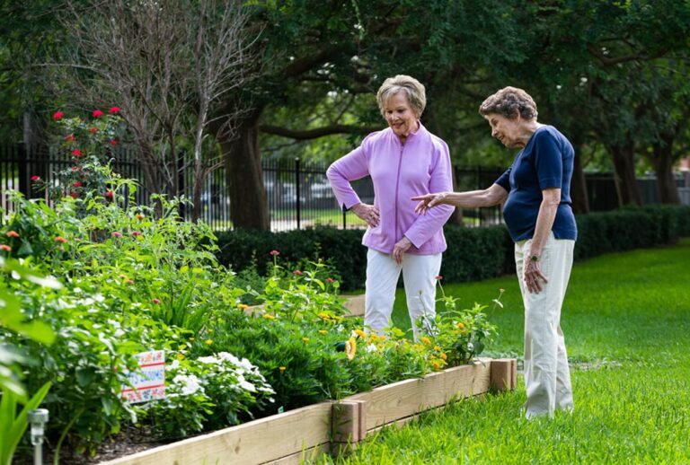 Two older women stand by a raised garden bed, looking at and discussing the flowers and plants growing in it.